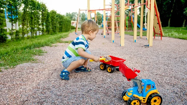 Ritratto di adorabile bambino di 3 anni che gioca con camion giocattolo con rimorchio nel parco giochi al parco. Bambino che scava e costruisce dalla sabbia — Foto Stock