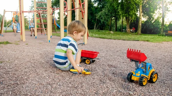 Portrait of adorable 3 years old toddler boy playing with toy truck with trailer on the playground at park. Child digging and building from sand — Stock Photo, Image