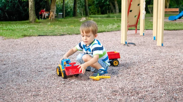 Portret cute 3 lat chłopiec maluch siedzi na placu zabaw w parku i gry z kolorowym plastikowym ciężarówką zabawka. Dziecko bawią się i bawią na zewnątrz z zabawkami — Zdjęcie stockowe