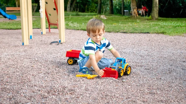 Closeup portrait of happy smiling 3 years old child boy digging sand on the playground with toy plastic truck or excavator. Child palying and having fun at park at summer — Stock Photo, Image