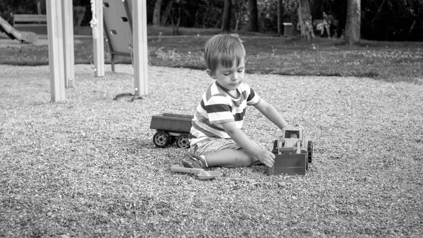 Black and white image of 3 years old toddler boy sitting in the sandbox at palyground and playing with toy truck excavator and trailer — Stock Photo, Image