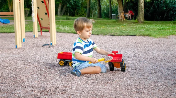 Porträt eines entzückenden 3-jährigen Kleinkindes, das auf dem Spielplatz am Park mit einem Spielzeug-LKW mit Anhänger spielt. Kind buddelt und baut aus Sand — Stockfoto