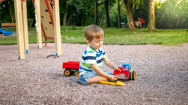 Immagine di primo piano di bambino carino che gioca sul palyground con i giocattoli. Bambino che si diverte con camion, escavatore e rimorchio. Sta fingendo di essere un costruttore o un autista — Foto Stock