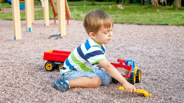 Porträtt av söta 3 år gamla småbarn pojke sitter på lekplatsen i parken och leker med färgglada plast leksaksbil. Barn ha roligt och leka utomhus med leksaker — Stockfoto