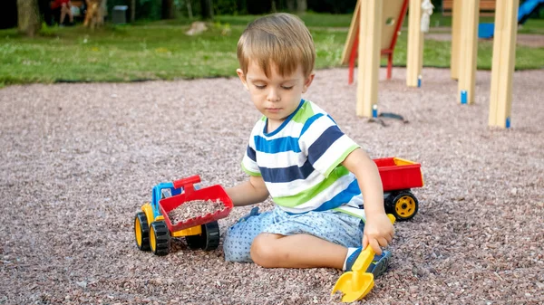 Närbild porträtt av Happy leende 3 år gammal barn pojke gräva sand på lekplatsen med leksaks plast lastbil eller grävmaskin. Barn palying och ha kul på Park på sommaren — Stockfoto