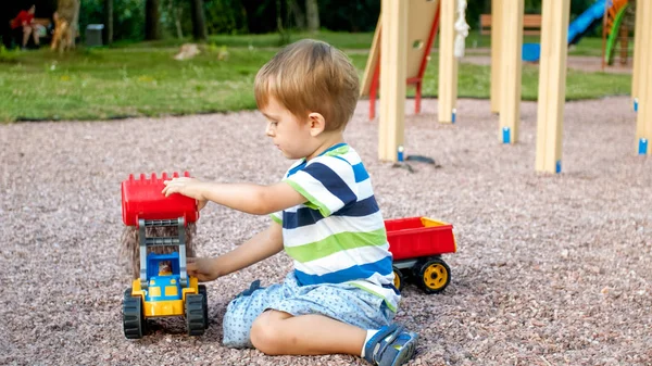 Retrato de adorável menino de 3 anos de idade brincando com caminhão de brinquedo com reboque no parque infantil no parque. Criança escavando e construindo a partir de areia — Fotografia de Stock