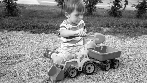Черно-белое изображение трехлетнего мальчика, сидящего в песочнице на палисаднике и играющего с игрушечным экскаватором грузовика и прицепом — стоковое фото