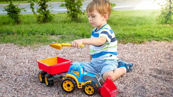 Retrato de un adorable niño de 3 años jugando con un camión de juguete con remolque en el parque infantil. Excavación y construcción de niños desde la arena — Foto de Stock