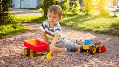 Fotoğraf sevimli 3 yaşındaki yürümeye başlayan çocuk kum ile oynarken ve kamyon ve römork parkta. Kum havuzunda çocuk kazma ve bina
