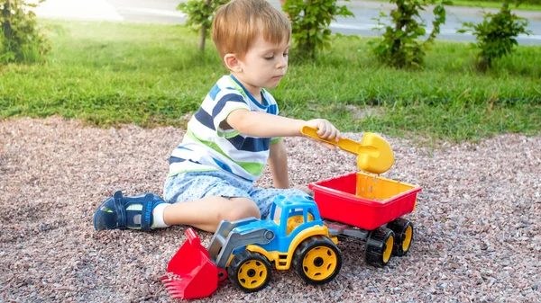 Portret cute 3 lat chłopiec maluch siedzi na placu zabaw w parku i gry z kolorowym plastikowym ciężarówką zabawka. Dziecko bawią się i bawią na zewnątrz z zabawkami — Zdjęcie stockowe
