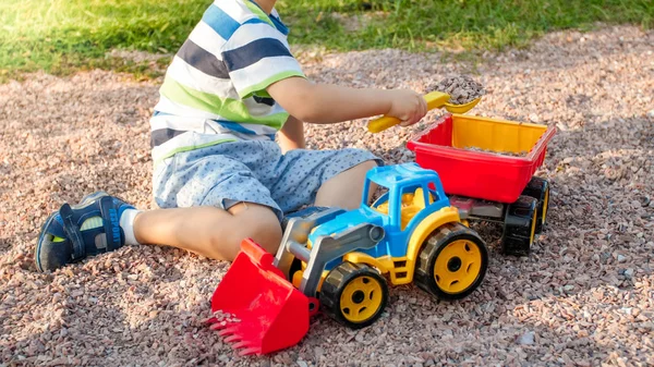 Primer plano retrato de feliz sonriente niño de 3 años cavando arena en el patio de recreo con camión de plástico de juguete o excavadora. Niño palying y divertirse en el parque en verano — Foto de Stock