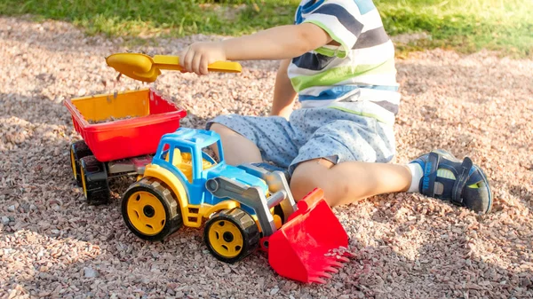 Ritratto di adorabile bambino di 3 anni che gioca con camion giocattolo con rimorchio nel parco giochi al parco. Bambino che scava e costruisce dalla sabbia — Foto Stock