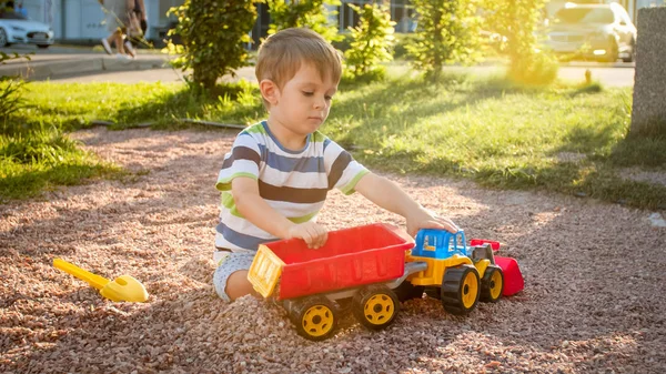 Κοντινό πορτρέτο του Happy χαμογελαστή 3 χρονών παιδί αγόρι σκάψιμο άμμου στην παιδική χαρά με πλαστικό φορτηγό παιχνίδι ή εκσκαφέας. Το παιδί να παλεύοντας και να διασκεδάζει στο πάρκο το καλοκαίρι — Φωτογραφία Αρχείου