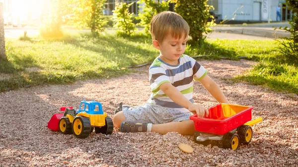 Immagine di primo piano di bambino carino che gioca sul palyground con i giocattoli. Bambino che si diverte con camion, escavatore e rimorchio. Sta fingendo di essere un costruttore o un autista — Foto Stock