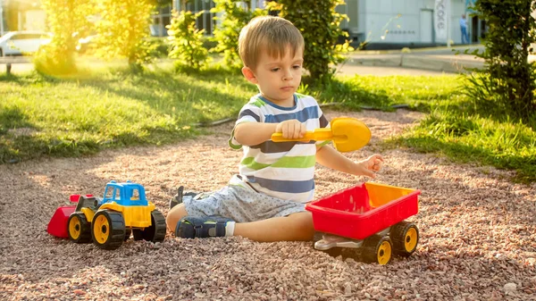 Immagine di primo piano di bambino carino che gioca sul palyground con i giocattoli. Bambino che si diverte con camion, escavatore e rimorchio. Sta fingendo di essere un costruttore o un autista — Foto Stock