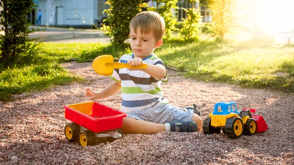 Portret cute 3 lat chłopiec maluch siedzi na placu zabaw w parku i gry z kolorowym plastikowym ciężarówką zabawka. Dziecko bawią się i bawią na zewnątrz z zabawkami — Zdjęcie stockowe