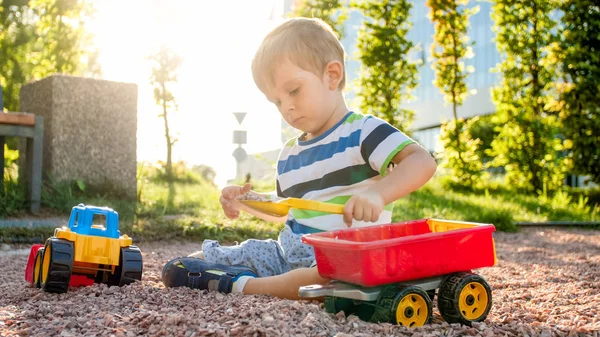 Närbild porträtt av Happy leende 3 år gammal barn pojke gräva sand på lekplatsen med leksaks plast lastbil eller grävmaskin. Barn palying och ha kul på Park på sommaren — Stockfoto