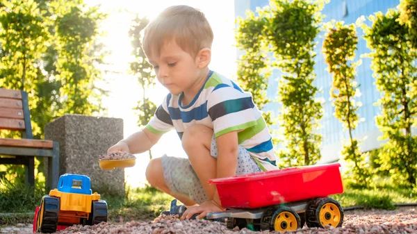 Photo of adorable 3 years old toddler boy playing with sand and you truck and trailer in park. Child digging and building in sandpit — Stock Photo, Image