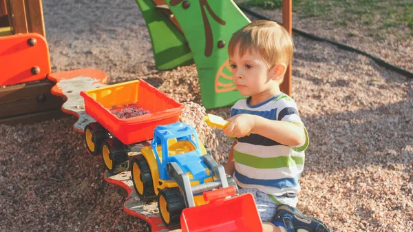 Retrato de niño sonriente sentado en la arena en el patio de recreo y cavando arena con pala de plástico y verterla en colorido camión de juguete con remolque —  Fotos de Stock