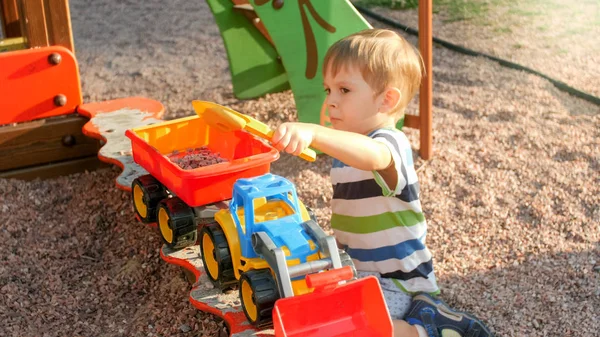 Portrait of cheerful smiling little boy pouring sand in toy truck with trailer. Kids playing and having on playground at park — Stock Photo, Image