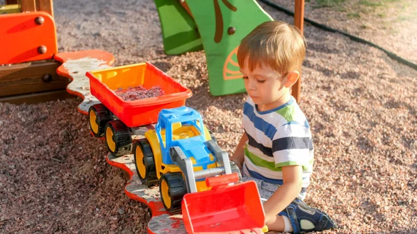Portret uśmiechniętego małego chłopca siedzącego w piaskownica na placu zabaw i kopania piasku z plastikową SPADE i wlewając go w kolorowe ciężarówki Zabawka z przyczepą — Zdjęcie stockowe