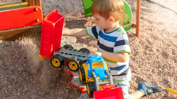 Retrato de niño sonriente alegre vertiendo arena en camión de juguete con remolque. Niños jugando y teniendo en parque infantil en el parque —  Fotos de Stock