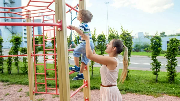Portriat młodej matki wspierając i trzymając jej 3-letni syn dziecka na drabinie metalowej na placu zabaw dla dzieci w parku — Zdjęcie stockowe