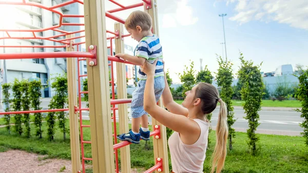 Zbliżenie wizerunek młodej matki pomagając jej mały syn wspinaczka na wysokich metalowych schodach na placu zabaw dla dzieci sportu — Zdjęcie stockowe