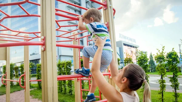 Portriat de jovem mãe apoiando e segurando seu filho de 3 anos de idade em escada de metal no parque infantil em parque — Fotografia de Stock