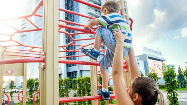 Close-up beeld van jonge moeder helpt haar kleine zoon klimmen op hoge metalen trappen bij sport kinderspeelplaats — Stockfoto