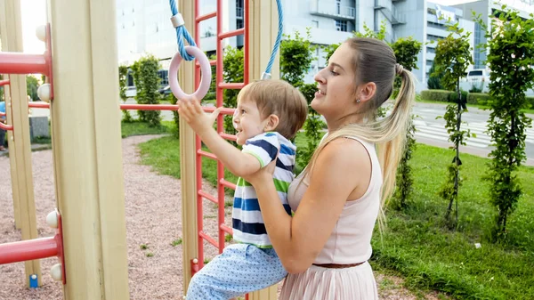 Portrait of cute smilling toddler boy hanging on the rings with his mother on children playground — Stock Photo, Image