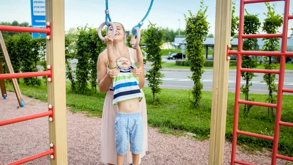 Portret van glimlachend 3 jaar oude kleine jongen doen pull-ups op ringen in sport speeltuin terwijl moeder houden en hem te suporteren — Stockfoto