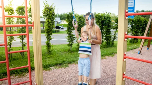 Portret van schattige smilling peuter jongen opknoping op de ringen met zijn moeder op kinderen speelplaats — Stockfoto