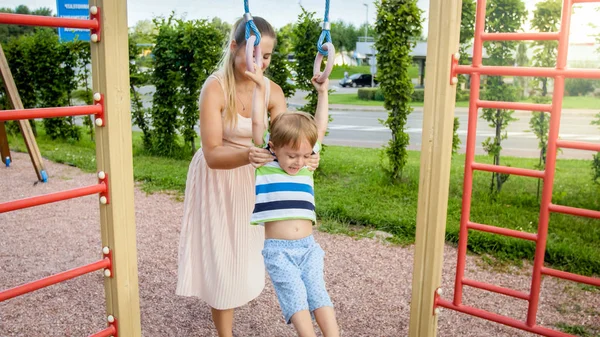 Close-up portret van jonge moeder te helpen en houden haar kleine zoon opknoping en zwaaien op sportrs ringen op palyground. Actieve kinderen die sporten — Stockfoto