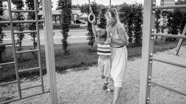 Retrato de un niño de 3 años sonriente haciendo flexiones en los anillos en el patio deportivo mientras la madre lo sostiene y lo apoya —  Fotos de Stock