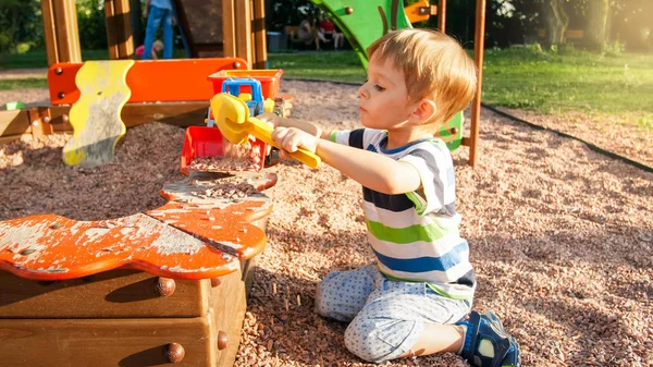 Porträtt av lilla 3 år gamla småbarn pojke sitter på lekplatsen och gräva sand med leksak plast spade — Stockfoto