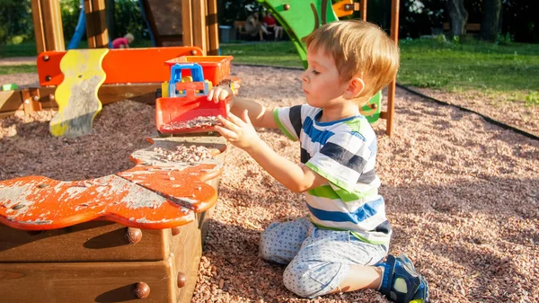 Portrait of smiling little boy sitting in the sandpit at playground and digging sand with plastic spade and pouring it in colorful toy truck with trailer — Stock Photo, Image