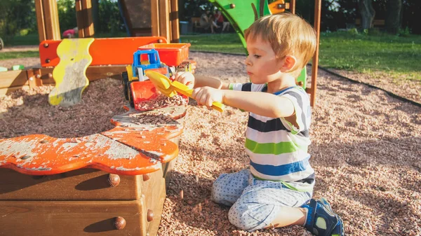 Portrait of smiling little boy sitting in the sandpit at playground and digging sand with plastic spade and pouring it in colorful toy truck with trailer — Stock Photo, Image