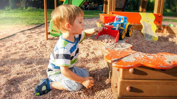 Portrait of little 3 years old toddler boy sitting on the playground and digging sand with toy plastic spade — Stock Photo, Image