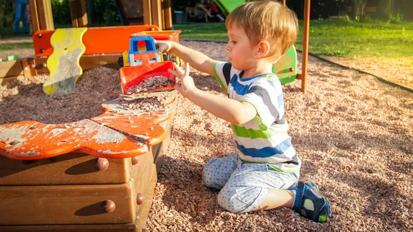 Porträt eines lächelnden kleinen Jungen, der im Sandkasten auf dem Spielplatz sitzt und mit einem Plastikspaten Sand ausgräbt und in einen bunten Spielzeugwagen mit Anhänger gießt — Stockfoto