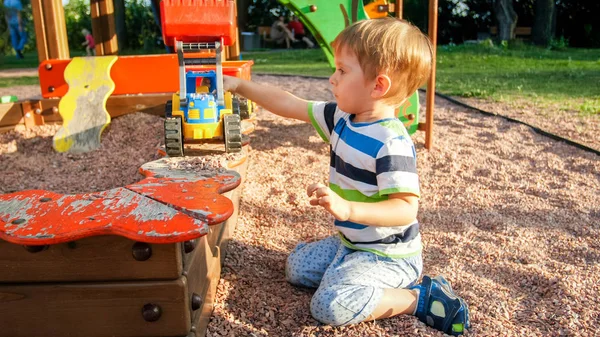 Porträtt av glad leende liten pojke hälla sand i leksaksbil med släpvagn. Barn leker och har på lekplats på Park — Stockfoto