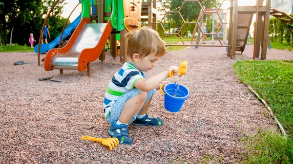 Photo of adorable 3 years old little boy sitting on the playground and digging sand with small plastic shovel and bucket — Stock Photo, Image