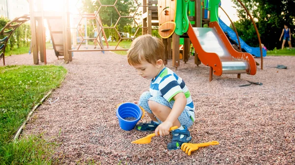 Foto eines entzückenden 3-jährigen Jungen, der auf dem Spielplatz sitzt und mit einer kleinen Plastikschaufel und einem Eimer Sand gräbt — Stockfoto