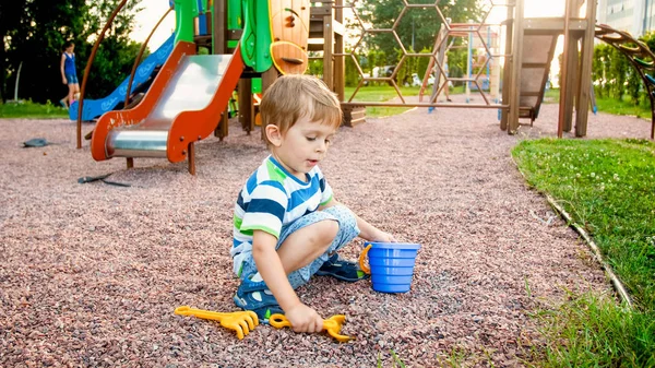 Imagem de menino sentado no parque infantil e puring areia com pequenas espadas de plástico em balde colorido. Kid escavação e construção no parque — Fotografia de Stock