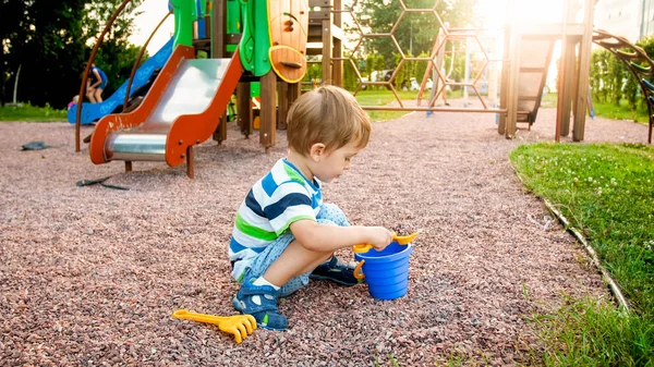Ritratto di un bambino di 3 anni seduto sul parco giochi e che scava sabbia con una pala di plastica giocattolo — Foto Stock