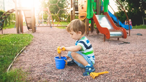 Image of little boy sitting on the playground and puring sand with small plastic spade in colorful bucket. Kid digging and building at park — Stock Photo, Image