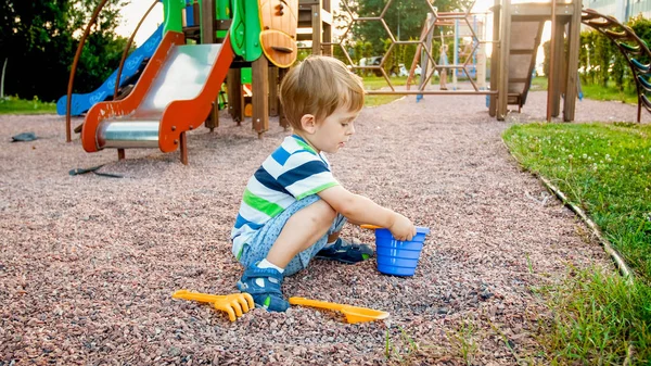 Foto eines entzückenden 3-jährigen Jungen, der auf dem Spielplatz sitzt und mit einer kleinen Plastikschaufel und einem Eimer Sand gräbt — Stockfoto