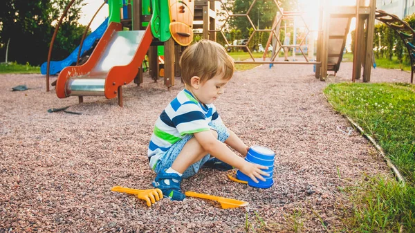 Image d'un petit garçon assis sur l'aire de jeux et purgeant du sable avec une petite bêche en plastique dans un seau coloré. Creuser et construire des enfants au parc — Photo