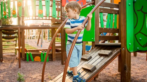 Portrait of active little boy crawling and climbing on wooden staircase on children playground at park — Stock Photo, Image