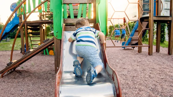 Foto de niño lindo de 3 años escalando y montando en un tobogán grande en el parque infantil — Foto de Stock